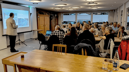 Informationsabend zum Thema künstliche Intelligenz im Restaurant Nollen. Referent Christian Blattner präsentiert vor über 30 Teilnehmenden, die an mehreren Tischen aufmerksam zuhören. Auf der Leinwand ist eine Präsentation zum Thema künstliche Intelligenz zu sehen.